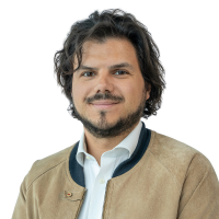 Portrait of a man with wavy dark hair and a light beard, wearing a tan jacket and light blue shirt against a white background.