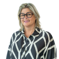 Portrait of a smiling woman with shoulder-length blond hair and glasses, wearing a black and white geometric-patterned blouse.