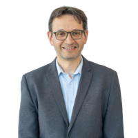 Head-and-shoulders portrait of a smiling man in a gray blazer and light blue shirt, wearing glasses.