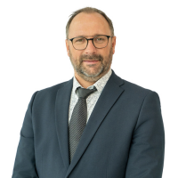 Professional headshot of a middle-aged man with short gray hair and beard, wearing a blue suit, tie, and glasses, against a white background.