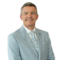 Portrait of a man wearing a light blue suit, white shirt, and patterned tie, smiling at the camera against a white background.