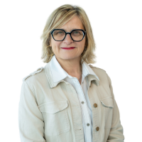Portrait of a blonde woman with black-framed glasses, wearing a light denim jacket and white shirt, looking at the camera with a gentle smile.